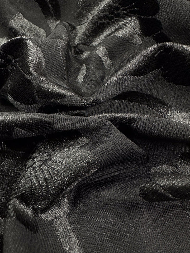 Super Cheap Fabrics' Burnout Velvet - Noir - 130cm features black burnout velvet with a floral pattern and shimmering highlights that catch the light. Its slightly crumpled texture adds sophistication and elegance to any design.