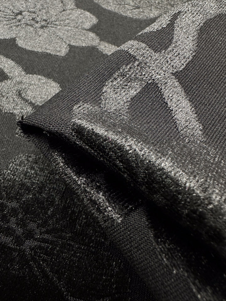 Close-up of two folded pieces of Burnout Velvet - Noir - 130cm by Super Cheap Fabrics, featuring a shiny, textured floral pattern. One piece is slightly rolled to reveal the intricate design. The lighting highlights the fabric's sheen and details.