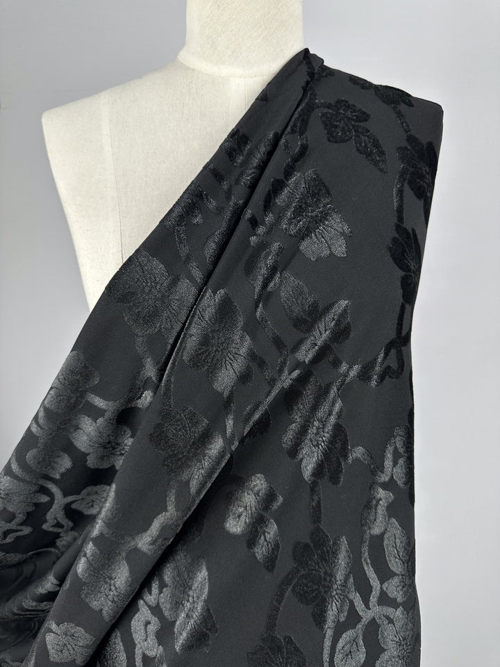 A mannequin showcases Super Cheap Fabrics' "Burnout Velvet - Noir - 130cm," featuring a luxurious floral pattern with a subtle, glossy finish that elegantly contrasts against a plain gray background.