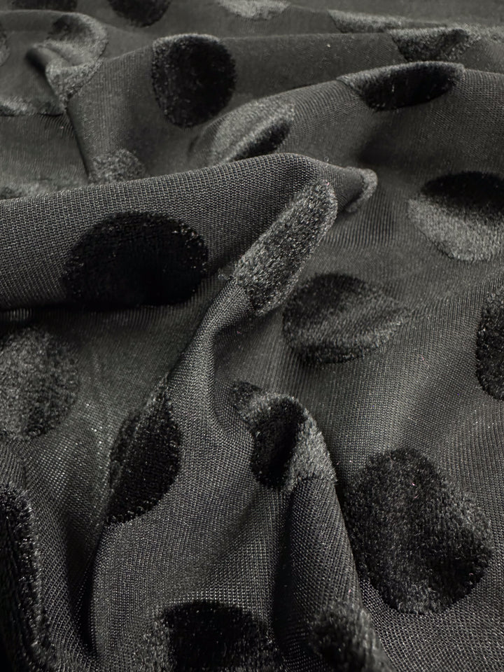 A close-up of Super Cheap Fabrics' Burnout Velvet - Polka Black, 155cm, showcases its luxurious fabric with large velvet polka dots. The cloth drapes softly, emphasizing the striking contrast between smooth texture and raised, fuzzy dots.