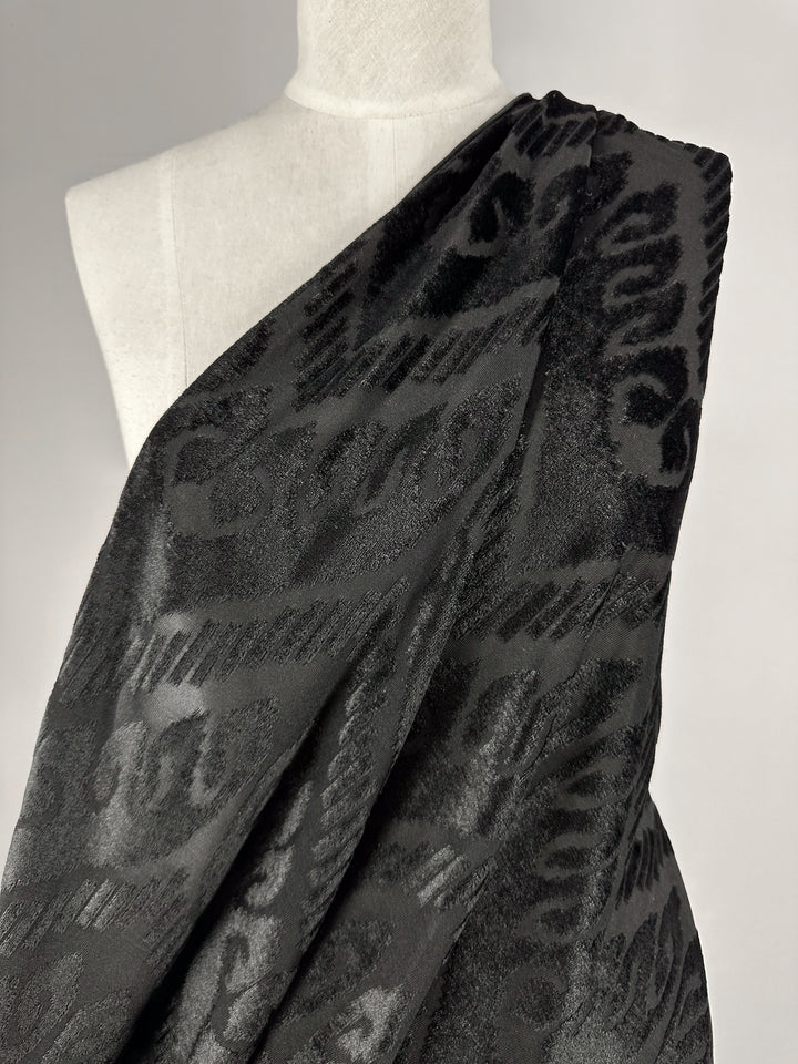 A mannequin showcases the Burnout Velvet - Arrows - 155cm fabric by Super Cheap Fabrics, exuding luxury with its intricate jacquard patterns and elegant sheen, reminiscent of an evening gown draped across the shoulder against a plain backdrop.