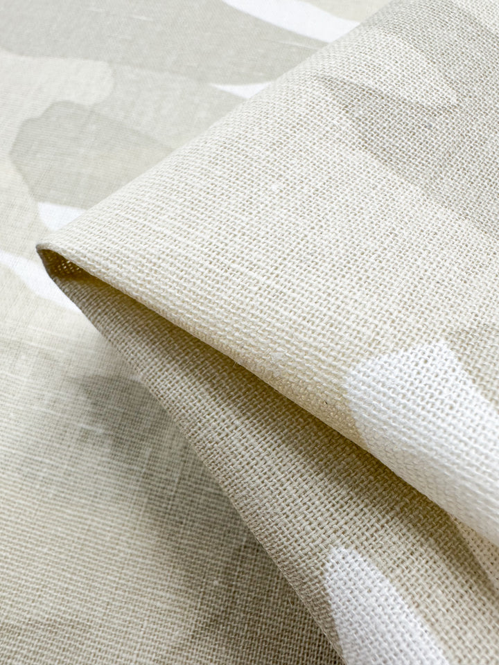 A close-up of a folded piece of Super Cheap Fabrics' Linen Blend - Calm Camo, 137cm wide, shows beige textured fabric with cotton. It features a subtle white abstract pattern and a natural woven look ideal for home decor, emphasizing its detailed weave and fibers.