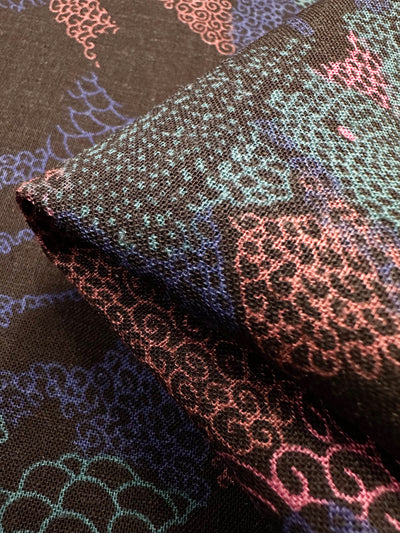 Close-up of folded Linen Blend - Coral - 147cm by Super Cheap Fabrics, featuring intricate pink, blue, and green patterns on a black background with swirling and scale-like motifs in a tapestry-like texture.