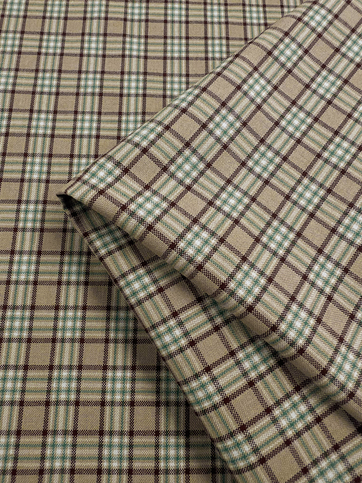 A folded view of the Shirting - Safari fabric by Super Cheap Fabrics, showing its beige and green plaid pattern on a complementary background. The design includes intersecting green, brown, and white lines crafted from a polyester cotton blend. This light to medium weight fabric is ideal for children's clothing due to its timeless tartan style.