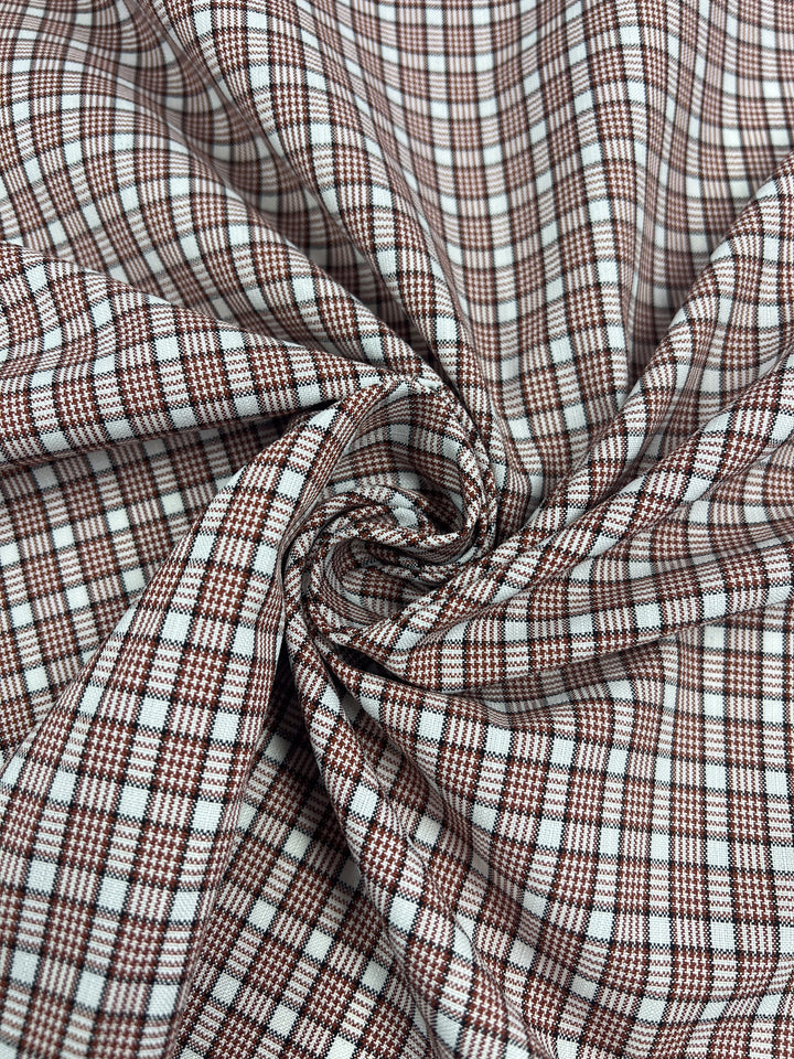 A close-up of "Shirting - Terra - 147cm" by Super Cheap Fabrics, showcasing brown and white plaid polyester cotton in a spiral. This lightweight fabric features a grid design with intersecting lines forming terracotta-tinged squares.