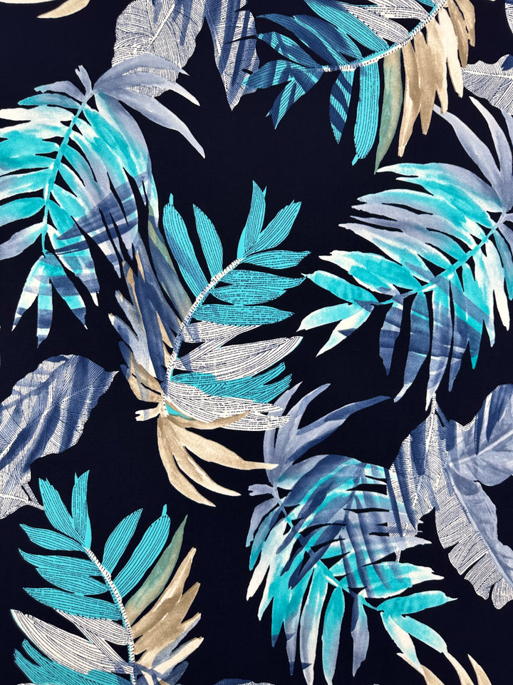 The Frost Tropic from Super Cheap Fabrics features blue, turquoise, and beige tropical palm leaves on a dark backdrop. This dynamic pattern on printed Lycra boasts a vibrant layered design and is 150cm wide.