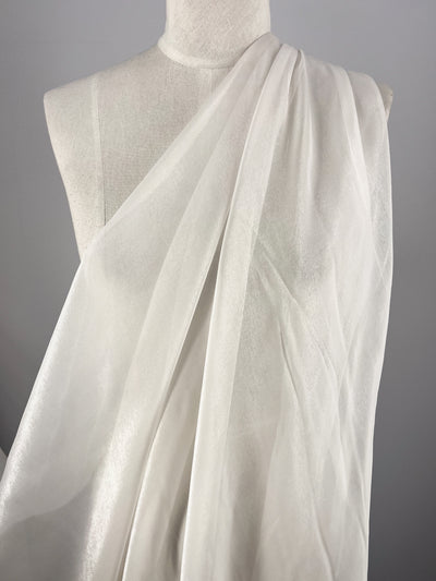 The Silk Mousseline - Bone by Super Cheap Fabrics is gracefully draped over a mannequin against a gray backdrop. This sheer, white fabric with its metallic shimmer beautifully reflects light, producing gentle variations in texture.