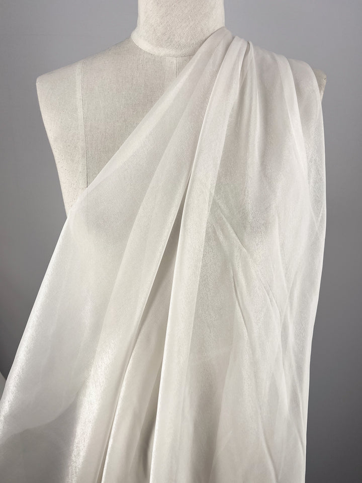 The Silk Mousseline - Bone by Super Cheap Fabrics is gracefully draped over a mannequin against a gray backdrop. This sheer, white fabric with its metallic shimmer beautifully reflects light, producing gentle variations in texture.