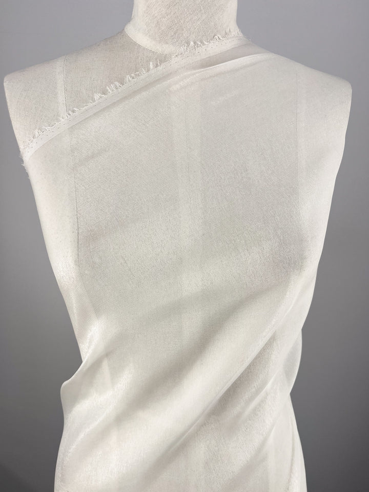 A piece of Silk Mousseline - Bone - 135cm from Super Cheap Fabrics is elegantly wrapped around a white dress form. The ultra-lightweight, sheer fabric exudes a subtle metallic shimmer, with its frayed edges cascading gracefully against the plain gray background.