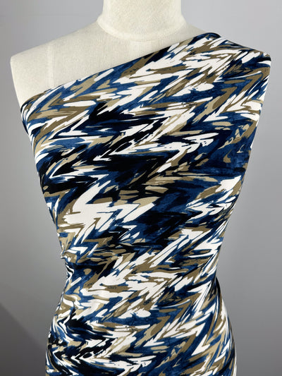 A mannequin wears a one-shoulder dress in Super Cheap Fabrics' Printed Lycra - Arrows, crafted from medium-weight Polyester/Spandex. The fabric boasts an abstract zigzag pattern in shades of blue, beige, black, and white against a neutral background for a dynamic visual effect.