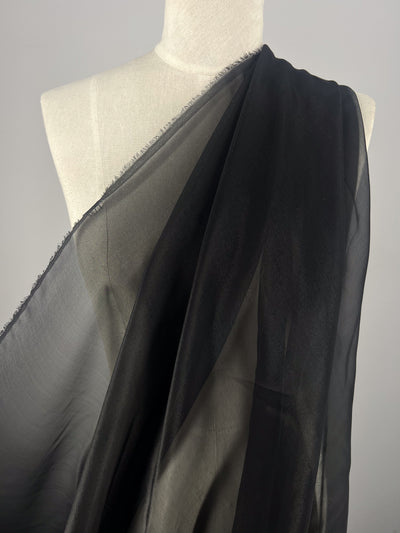 A mannequin displays Super Cheap Fabrics' Silk Mousseline in black, highlighting its sheer texture and transparency with a metallic shimmer overlay draped over the shoulder, flowing elegantly against a plain neutral background.