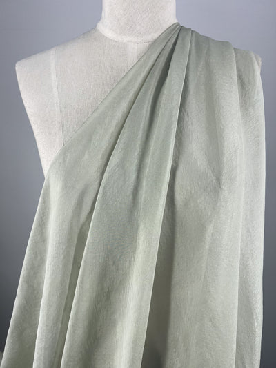 A mannequin displays Super Cheap Fabrics' Silk Mousseline - Frost, draped with soft pale green fabric infused with a metallic shimmer over one shoulder. This 140cm silk is light and airy, slightly textured, set against a neutral gray background.