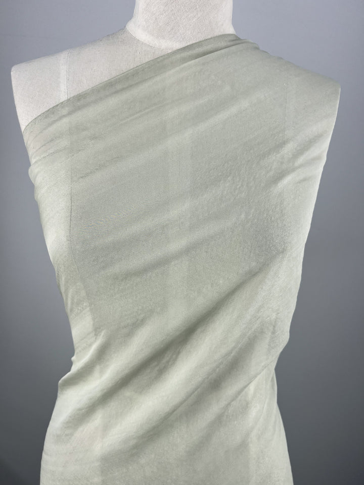 A mannequin displays the Silk Mousseline - Frost - 140cm by Super Cheap Fabrics, showcasing its texture and translucency. The semi-sheer, light gray silk is wrapped asymmetrically over one shoulder, its metallic shimmer enhancing its fluidity against a gray backdrop.