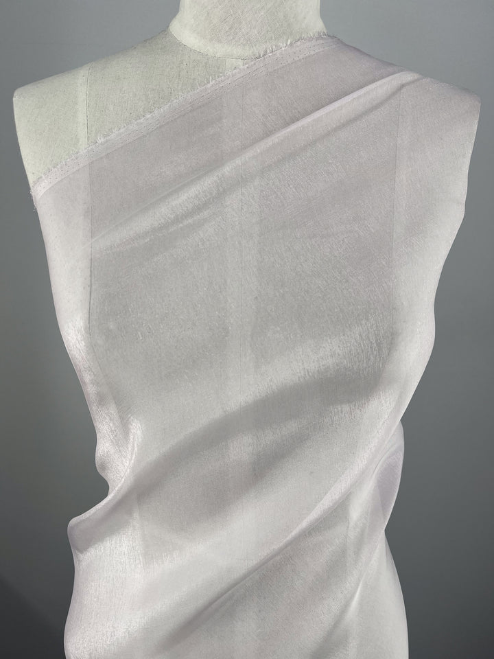 The Silk Mousseline - Orchid by Super Cheap Fabrics gracefully drapes over a mannequin, showcasing its sheer, lightweight texture with a subtle metallic shimmer. Its elegant translucence and fluid lines shine against a neutral gray backdrop.