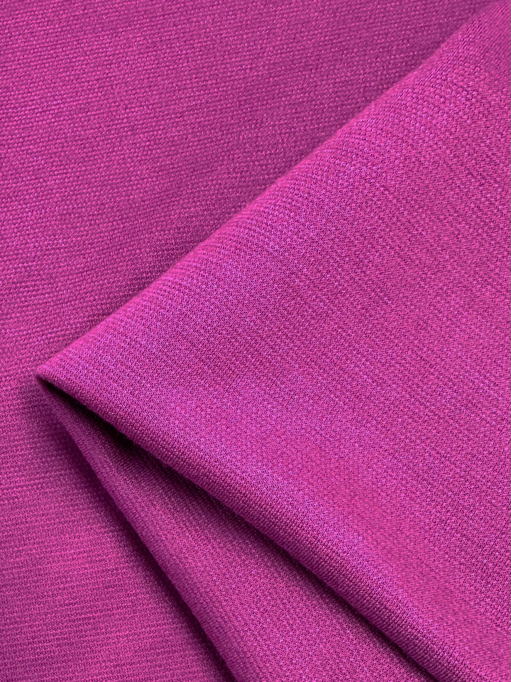 A close-up view of the vibrant fuchsia Waffle Knit fabric from Super Cheap Fabrics reveals its intricate honeycomb pattern and slightly textured, smooth surface. The fabric is folded over itself, showcasing its pliability and substantial thickness. The three-dimensional effect of the 152cm wide knit adds depth, creating a bold and striking visual.