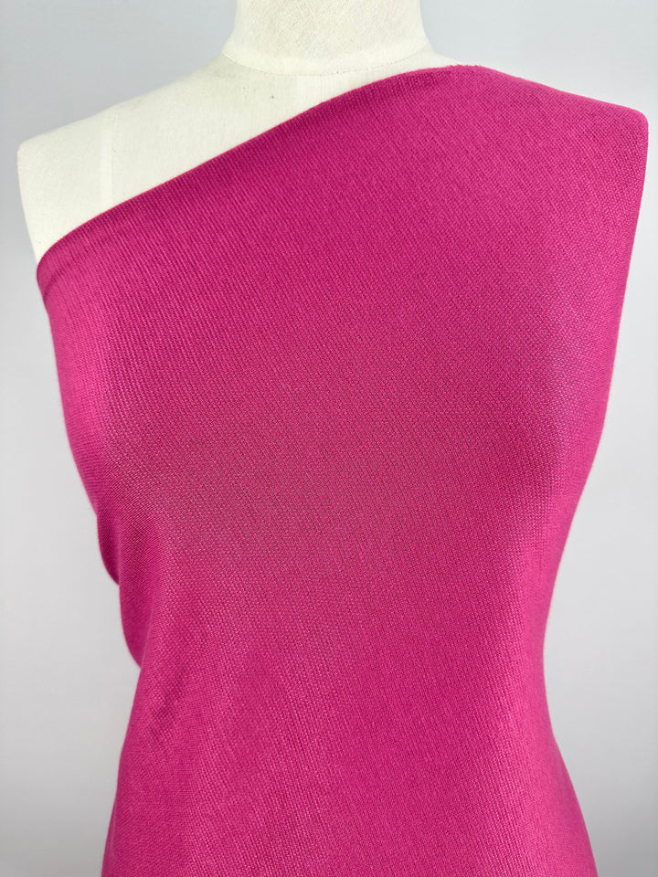 A mannequin is dressed in a vibrant fuchsia, one-shoulder knit garment made from the Waffle Knit - Fuchsia - 152cm fabric by Super Cheap Fabrics. The smooth-textured material features a subtle three-dimensional effect, and the asymmetrical design leaves one shoulder exposed. The plain, neutral background ensures that all attention remains on the striking garment.