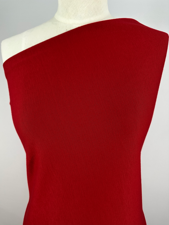 A mannequin is adorned with a vibrant red, off-shoulder top crafted from the "Waffle Knit - Fire - 152cm" fabric by Super Cheap Fabrics against a plain background. The soft, textured material lends an elegant touch and provides a subtle three-dimensional appearance.