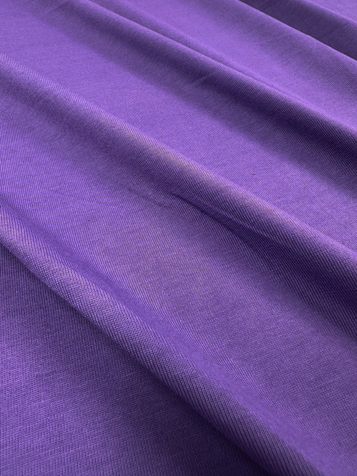 A vibrant Patrician Purple cotton fabric from Super Cheap Fabrics drapes smoothly, highlighting its rich color and soft texture. The lighting enhances the elegance of this 170cm medium-weight Cotton Jersey, showcasing gentle folds.