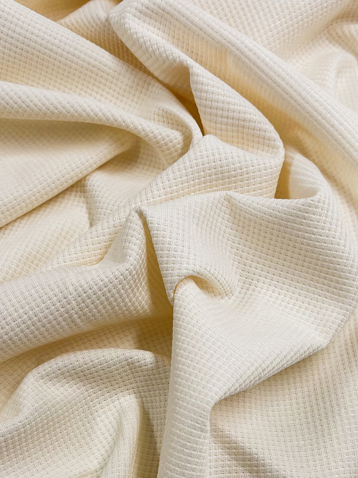 A close-up of the Super Cheap Fabrics' Waffle Knit in cream, featuring a delicate grid texture. The fabric's soft, crumpled folds craft an intricate and three-dimensional interplay of light and shadow. Measuring 170 cm wide, it showcases a rich design that brings out its luxurious texture.