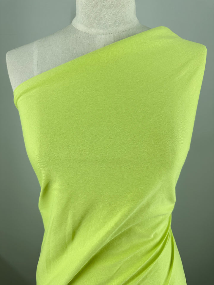 A mannequin is adorned with Super Cheap Fabrics' Cotton Jersey in Daiquiri Green (170cm), styled into a one-shoulder design. The smooth material, with subtle folds, highlights its texture and vibrant color against a soft, blurred gray-green background.