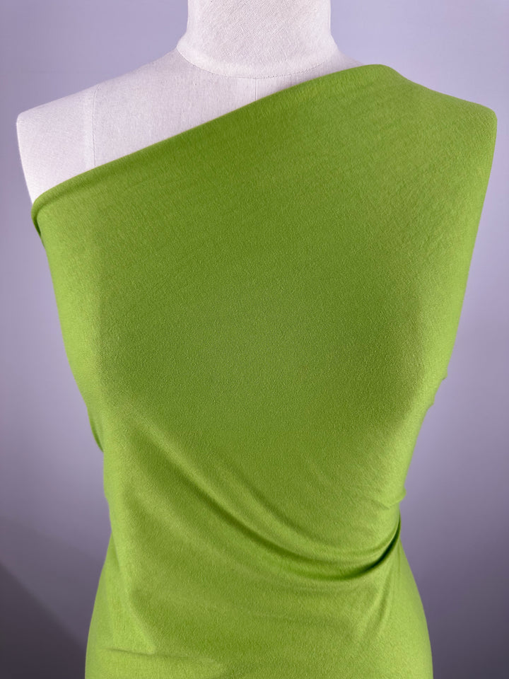 A mannequin showcases a chic, one-shoulder garment in lime green Cotton Jersey - Titanite by Super Cheap Fabrics. The smooth, slightly fitted fabric complements the contemporary design against a plain gray background.