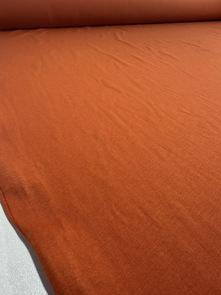 A large roll of Cotton Jersey in apricot orange by Super Cheap Fabrics is gently stretched on a flat surface, displaying its soft texture and subtle creases. The rich color adds warmth and highlights its versatile lightweight quality.