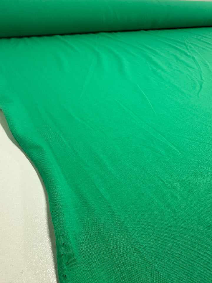 A close-up shows the Cotton Jersey in Vivid Green from Super Cheap Fabrics, partially unrolled on a flat surface. The 165cm fabric displays a smooth texture with visible creases and folds.