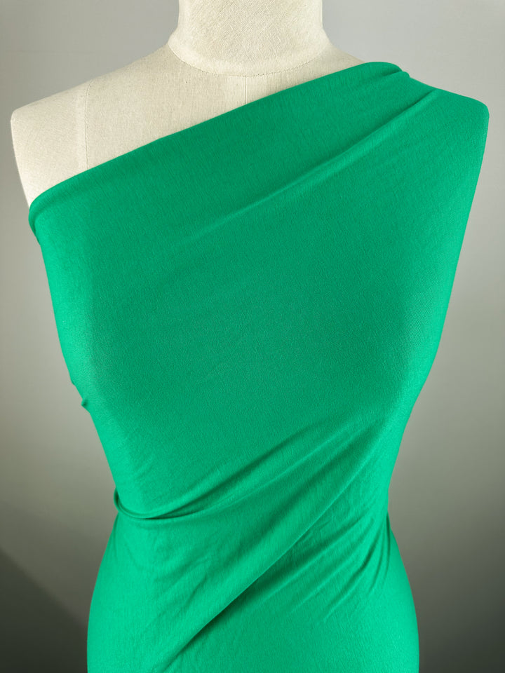 A mannequin draped in Super Cheap Fabrics' Vivid Green Cotton Jersey (165cm) showcases a one-shoulder garment with a smooth, flowing texture against a neutral background.