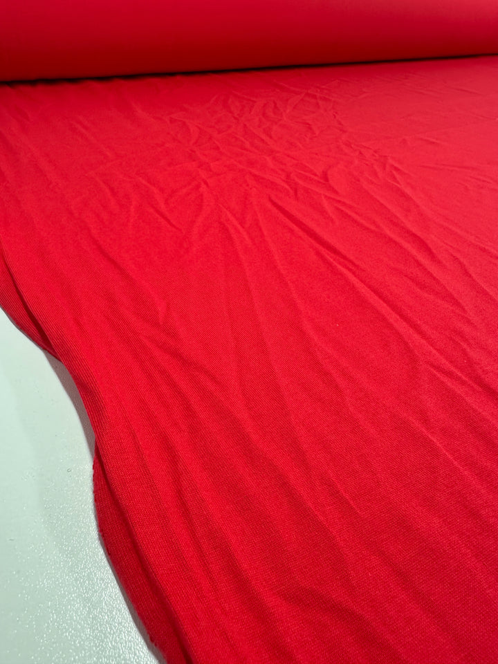 A close-up of the Super Cheap Fabrics' Cotton Jersey - Ribbon Red 165cm partially unrolled on a white surface shows its smooth texture and slight creases, indicating a soft, flexible medium weight jersey.