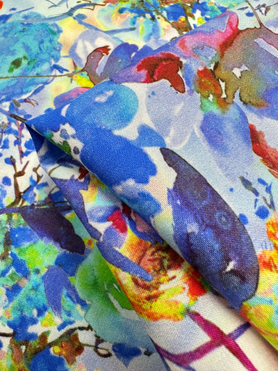 A close-up of Designer Rayon - Cortney by Super Cheap Fabrics reveals a vibrant abstract pattern with splashes of blue, red, yellow, and green like a watercolor painting. The 145cm fabric drapes beautifully to show its texture and dynamic prints.