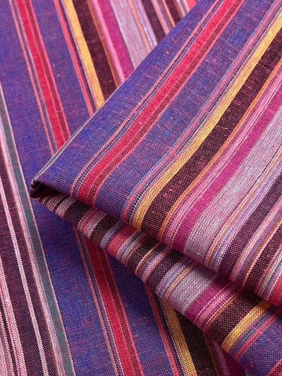 Close-up of the Linen Blend - Goa - 150cm by Super Cheap Fabrics, showcasing a vibrant, multicolored striped fabric with shades of purple, pink, yellow, and red. The yarn-dyed fabric is folded to highlight its texture and alternating stripe patterns for a vintage appearance.