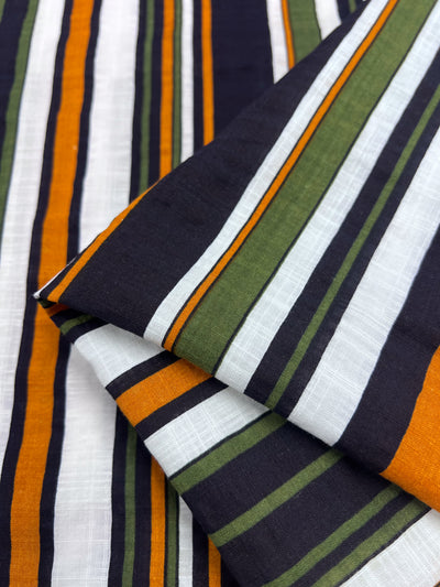 Close-up of the Linen Blend - Retro fabric, featuring breathable material with vibrant vertical stripes in green, orange, white, and black. This Super Cheap Fabrics product showcases an overlapping pattern that creates a dynamic and textured visual effect, making it perfect for summer outfits.