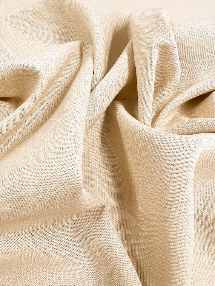 Close-up of the Premium Linen Cotton in a shortbread hue, offered by Super Cheap Fabrics. The slightly textured surface fabric is lightweight and measures 140cm. It is gently gathered and draped, creating subtle folds and shadows as the light enhances its warm and cozy appearance.