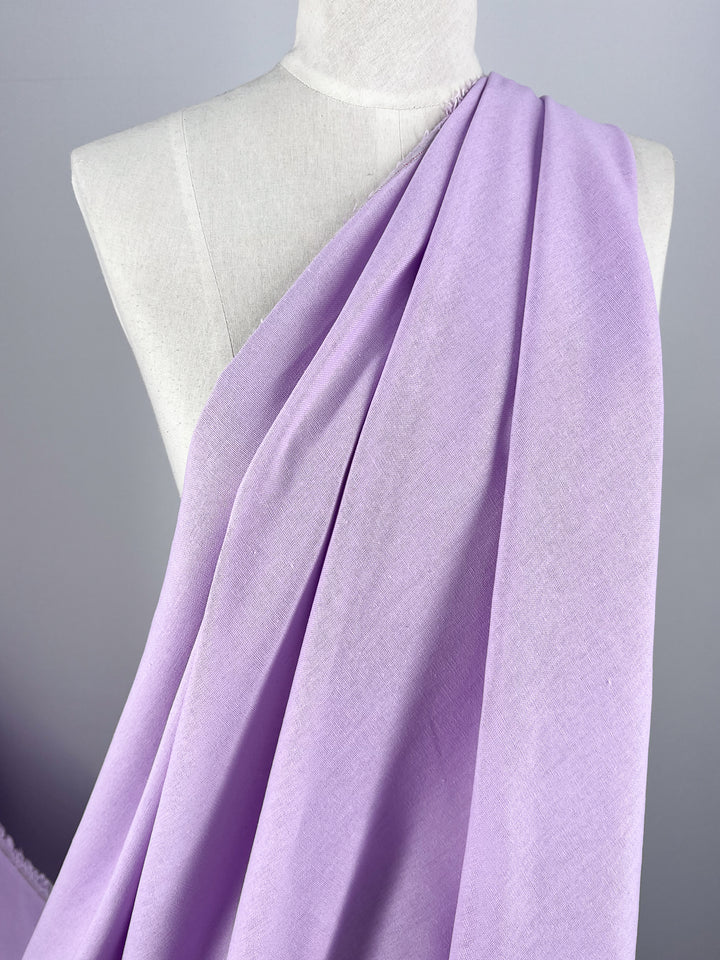 A mannequin is elegantly adorned with Super Cheap Fabrics' Premium Linen Cotton in "Orchid Bouquet," a lightweight, light purple fabric. Draped diagonally across the torso, the fabric showcases its soft texture and gentle folds against a plain gray background, highlighting its exquisite beauty.