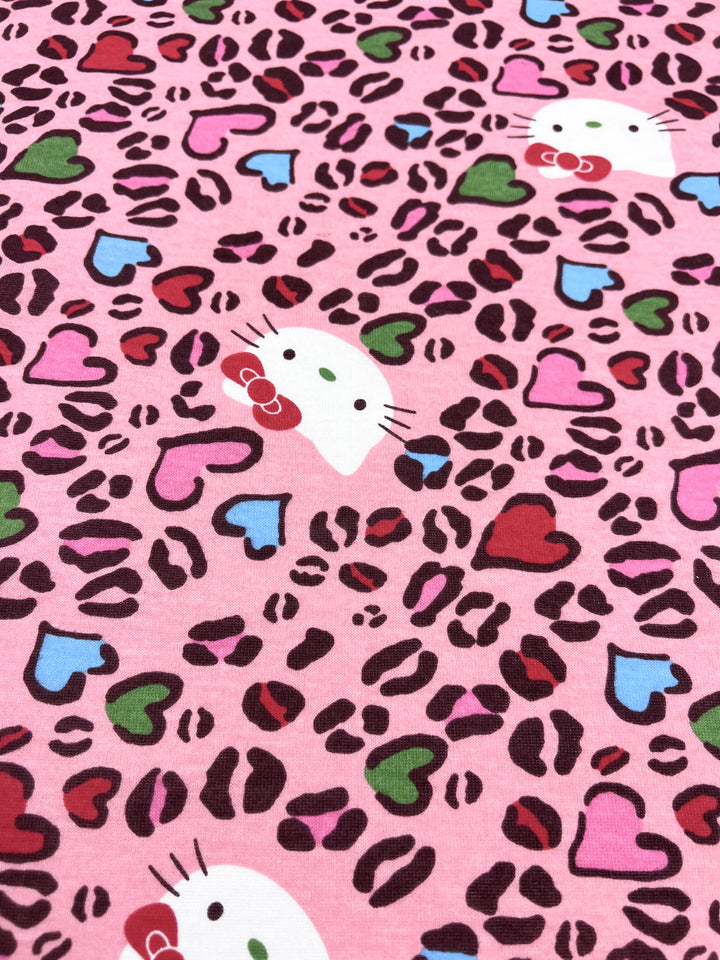 Introducing "Hello" by Super Cheap Fabrics, a lightweight printed jersey fabric measuring 150cm. This whimsical design features charming pink cartoon cat faces adorned with bows, interspersed with playful leopard print spots and vivid heart shapes in red, pink, blue, and green.