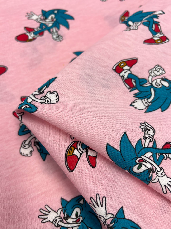 This lightweight printed jersey fabric from Super Cheap Fabrics features a vibrant pink background with animated Sonic characters in dynamic action poses, complete with their iconic red shoes and white gloves. Made from a soft poly/cotton jersey blend, this material measures 165cm in width.