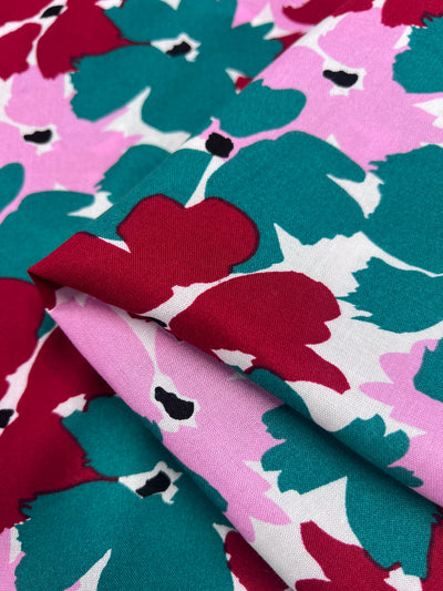 A close-up of Designer Rayon - Spring by Super Cheap Fabrics displays a vibrant floral pattern in shades of red, pink, teal, and white. The folded fabric reveals abstract, overlapping flower shapes in its colorful design.