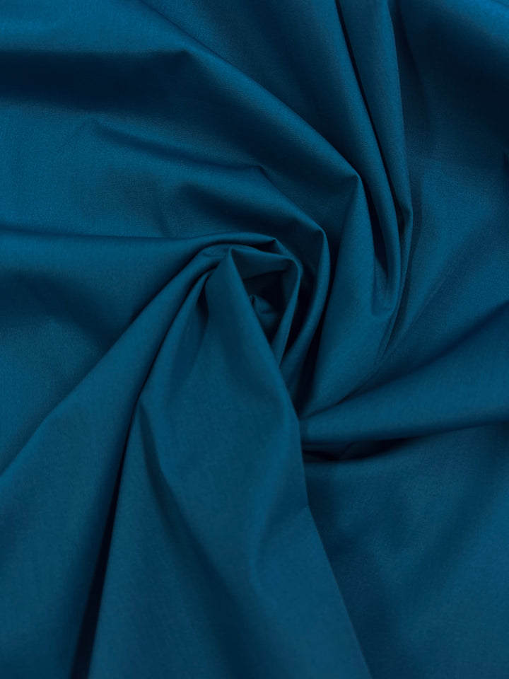 A close-up of crumpled Poplin - Celestial from Super Cheap Fabrics, a soft poly/cotton blend in dark teal with a central spiral pattern, shows its inviting texture and varying shades due to the folds—ideal for beginners exploring textiles.