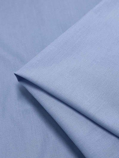 A close-up of Super Cheap Fabrics' "Oxford Poplin - Vista Blue - 148cm," a light blue polyester-cotton blend, displays its smooth, slightly textured surface. The non-stretch material is folded neatly, highlighting its soft texture and subtle sheen.