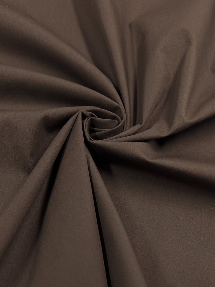 A close-up of Super Cheap Fabrics' Poplin - Rocky Road, a 148cm wide brown polyester cotton blend, swirled in a spiral. The lightweight fabric is smooth with a matte finish and soft folds, perfect for childrenswear.