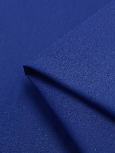 A folded piece of Super Cheap Fabrics' Poplin - Bluing showcases dark blue polyester cotton blend with a smooth texture. The 148cm non-stretch, lightweight fabric is creased at the corner, forming a geometric shape and possessing a subtle sheen.