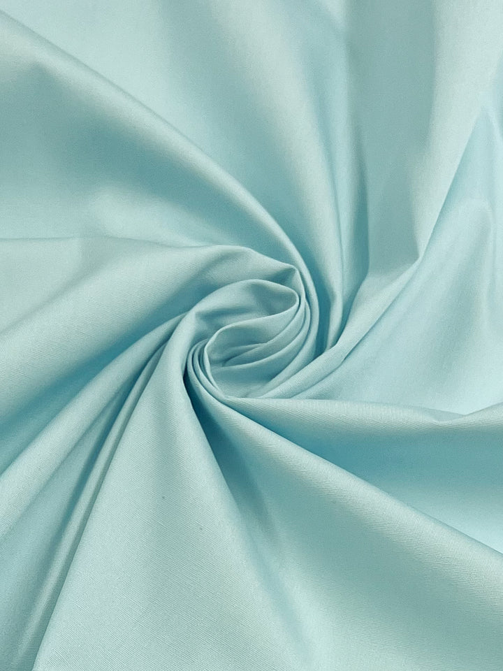 A close-up of Super Cheap Fabrics' Poplin - Cooling Spray, 148cm wide, reveals light blue polyester cotton blend artistically arranged in a spiral. Its smooth texture and gentle folds create a soft appearance ideal for showcasing fluidity and subtle sheen in quality childrenswear.