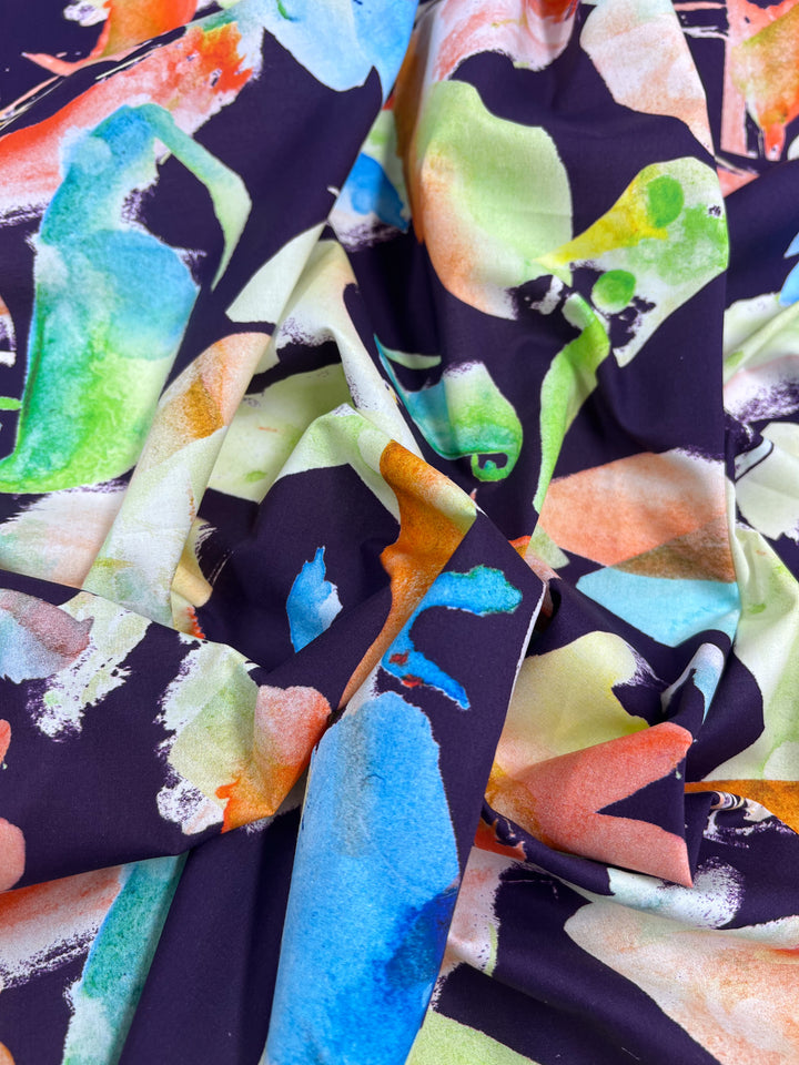 A close-up of Super Cheap Fabrics' "Printed Cotton - Chiken - 150cm" reveals lightweight cotton with a vibrant abstract watercolor pattern in green, blue, orange, and yellow on a dark backdrop. The fabric's natural fibers offer a loose drape and elegant folds.