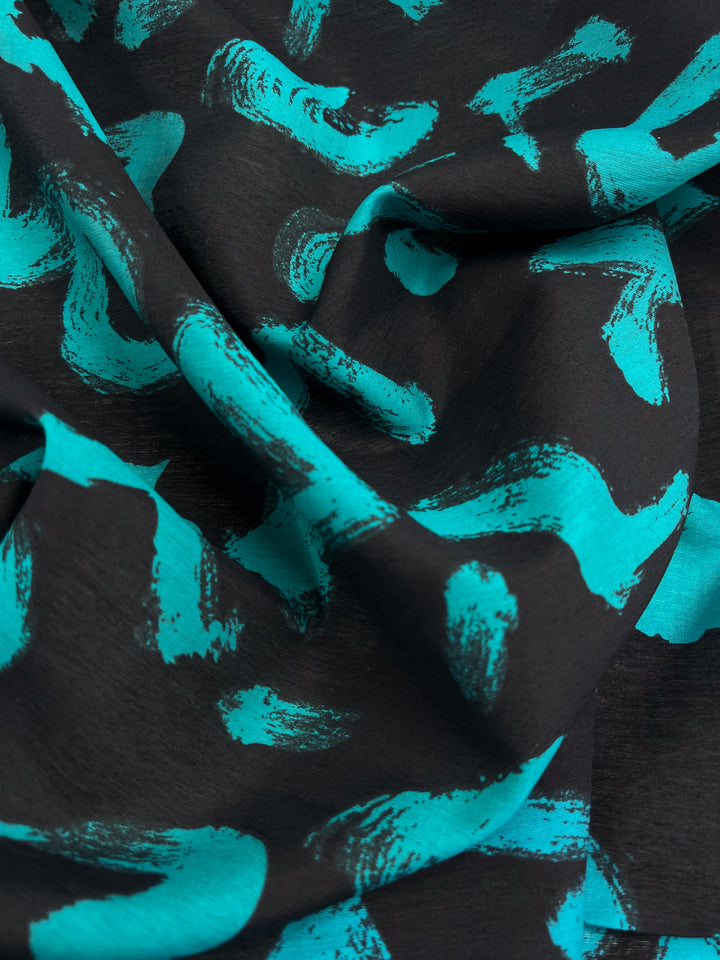 Close-up of a textured, slightly wrinkled black base fabric with turquoise abstract brushstrokes. The fluidity and intricate pattern are showcased on the 150cm "Printed Cotton - Texter" by Super Cheap Fabrics.