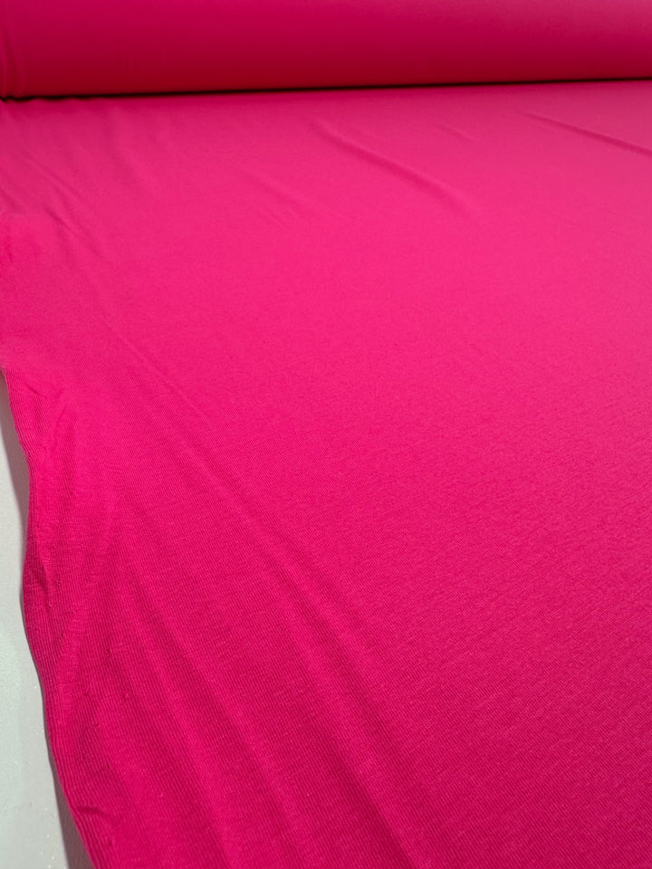 A large roll of Cotton Lycra - Fandango Pink fabric by Super Cheap Fabrics is unrolled on a white surface, showcasing its smooth texture and vibrant color. Ideal for crafting stunning bodycon dresses, its bold hue dominates the frame.