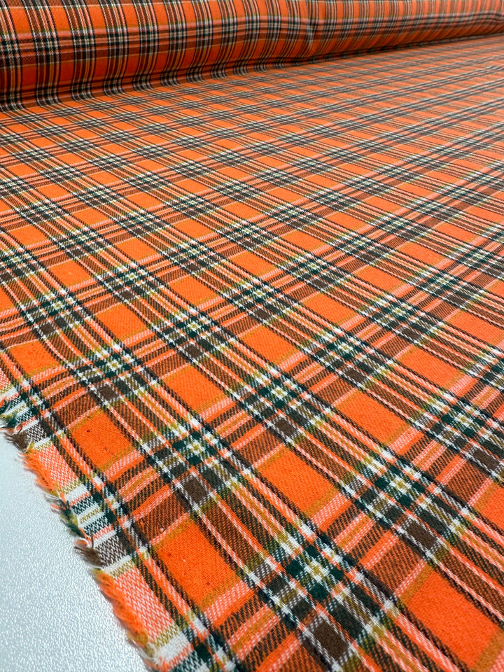 A close-up view of a roll of the **Suiting - Orange Glen Check - 147cm** fabric by **Super Cheap Fabrics**. The lightweight material is laid out on a flat surface, showcasing its intricate orange, green, white, and black tartan plaid pattern. The edges of this versatile fabric appear slightly frayed.