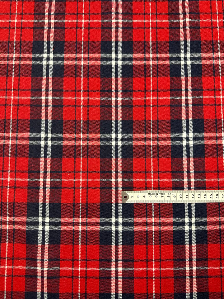 An up-close image showcases the "Suiting - Trad Tartan - 147cm" fabric by Super Cheap Fabrics, featuring a red plaid pattern with black, white, and dark red vertical and horizontal stripes. A ruler is positioned at the bottom-right corner to provide scale for the pattern size. This lightweight material is perfect for twill suiting or crafting formal suits.