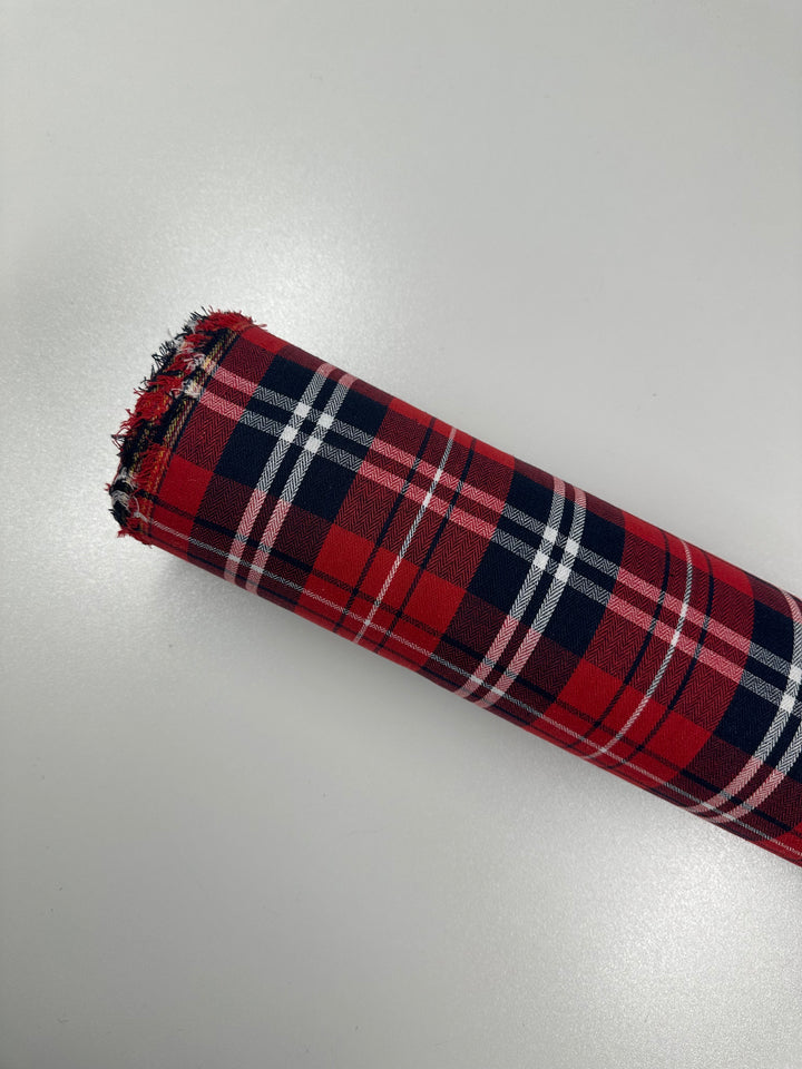 A roll of Super Cheap Fabrics' Suiting - Trad Tartan - 147cm, showcasing a lightweight twill suiting with a red, navy blue, white, and black tartan plaid pattern, is displayed against a plain, light-colored background. The fabric edges at the top of the roll show some fraying.