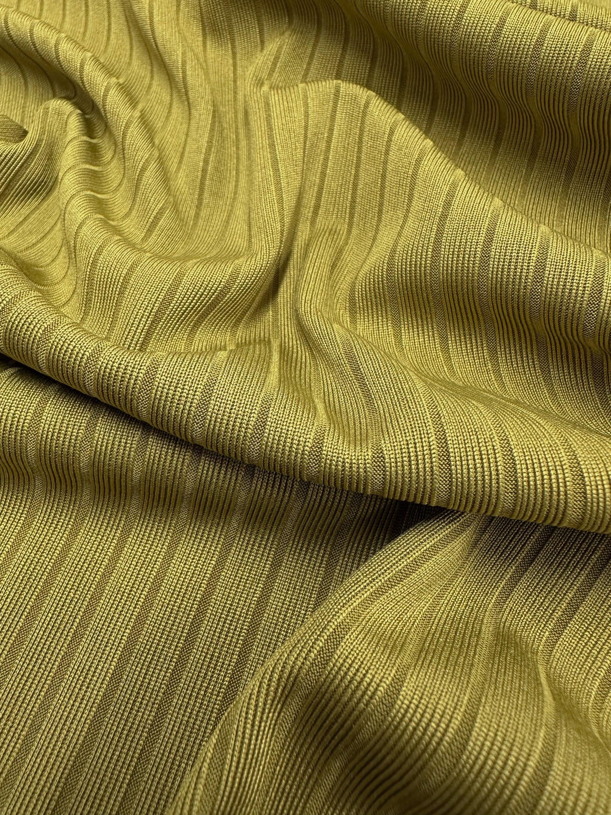 A close-up of Super Cheap Fabrics' Rib Knit - Avocado Oil, 173cm, showcases a textured yellowish-olive medium-weight knit with vertical lines for movement and depth, providing stretch and comfort.