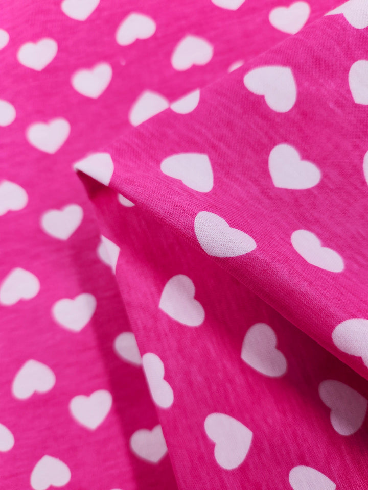 Close-up of Super Cheap Fabrics' Printed Jersey - Hearts - 160cm, featuring a bright pink base with white hearts. This medium-weight fabric is slightly crumpled, creating shadows and highlights for added texture depth.